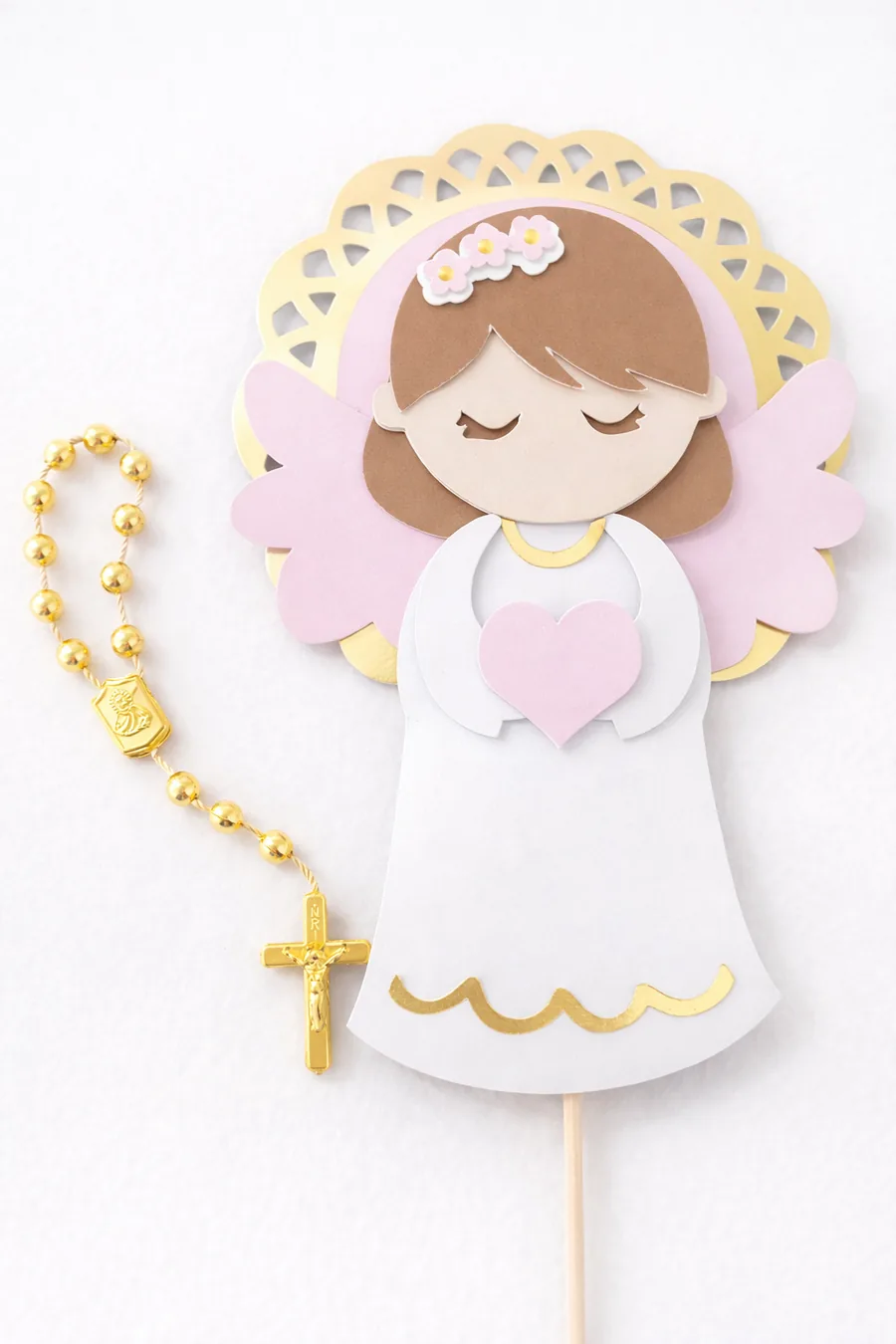 Topo de batizado com anjo em papel branco, asas cor-de-rosa e moldura dourada, ao lado de um terço