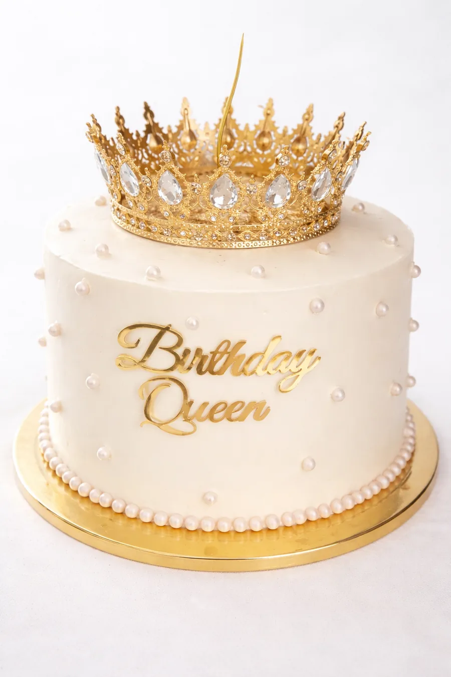 Bolo branco com pérolas, coroa dourada e a frase Birthday Queen recortada em dourado